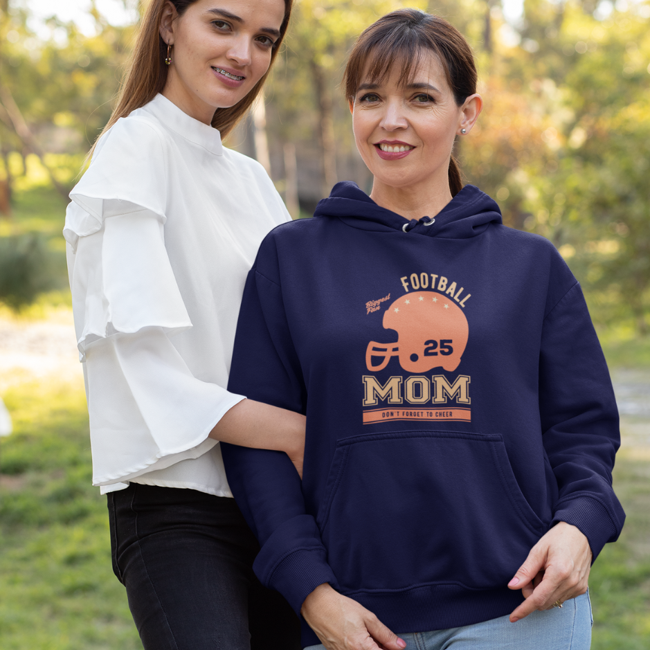 A mom wearing a navy hoodie with "Football Mom: Don't Forget to Cheer" text, standing with her young daughter wearing a white chemise in a garden.