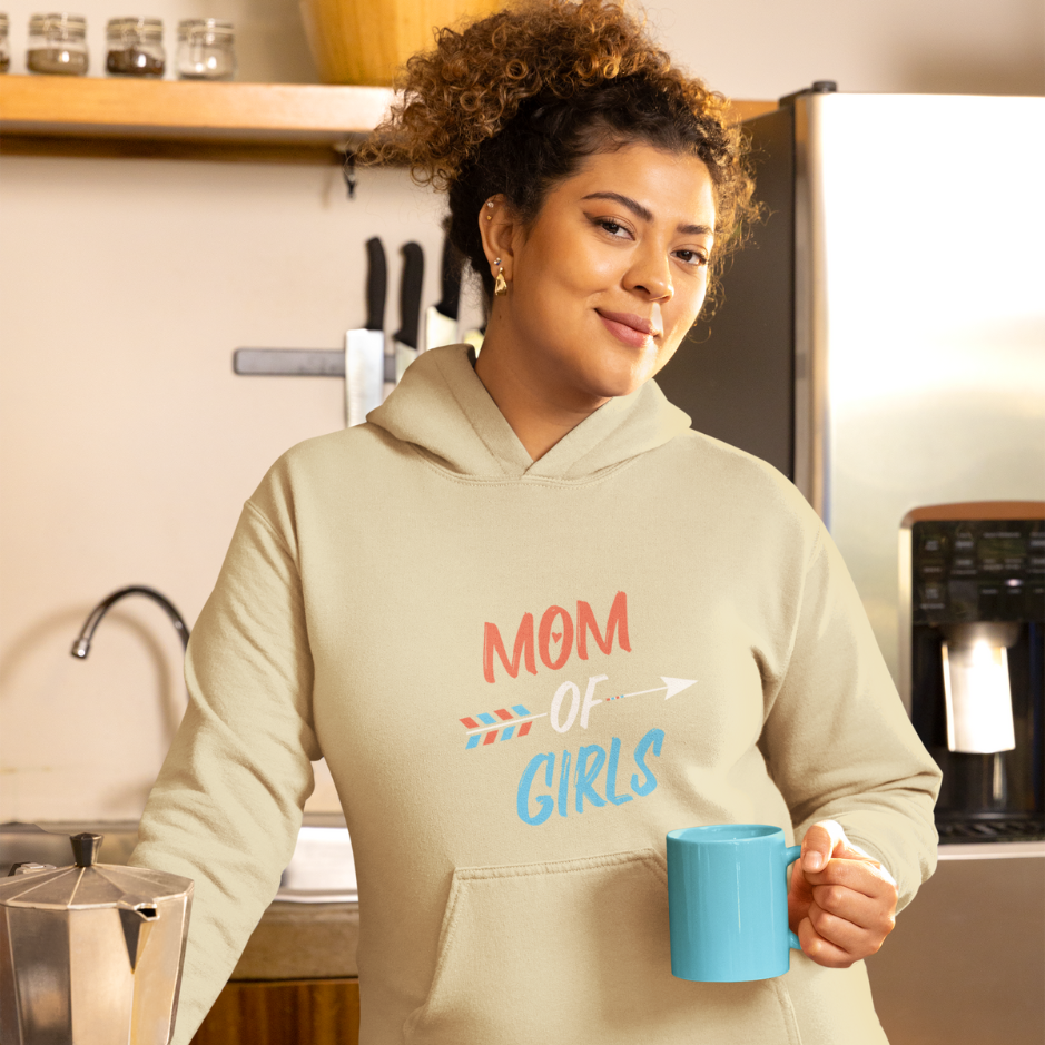 A pregnant mom stands in the kitchen, holding a blue cup, wearing a sand beige hoodie themed as 'Mom of Girls